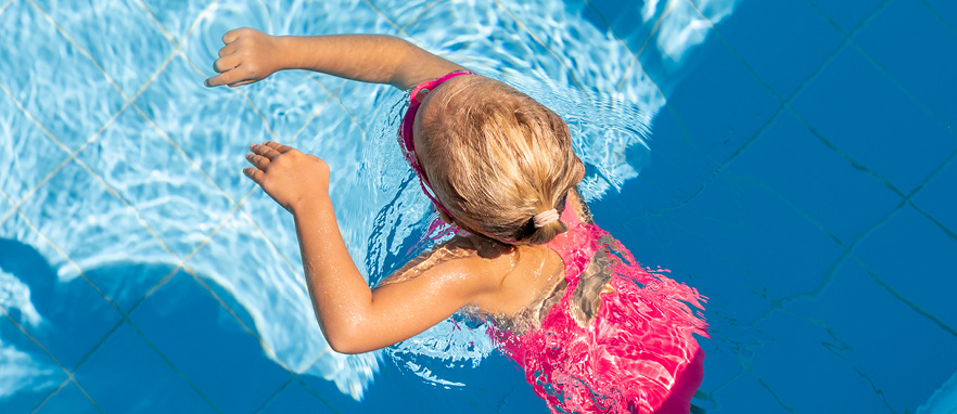 young girl in pink bathing suit and goggles splashing around in a light blue tiled swimming pool
