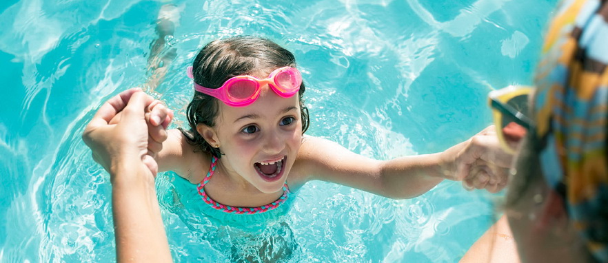 small girl with pink goggles in swimming pool holding a womans hands